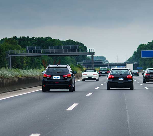 Several vehicles drving along a multi-lane highway with overhead road signs and trees.