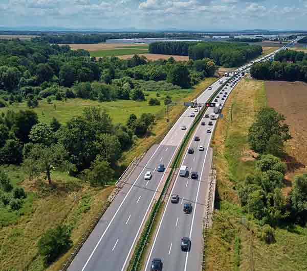 traffic moving a long a highway surrounded by tress and farmland.