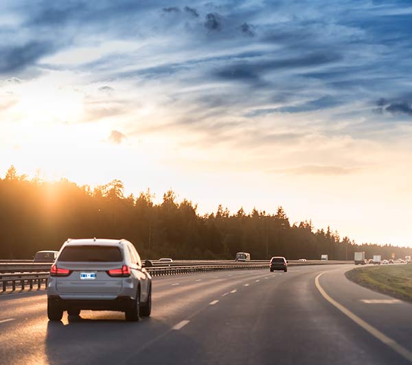 A white SUV driving along a curved highway at sunset with trees lining the road.